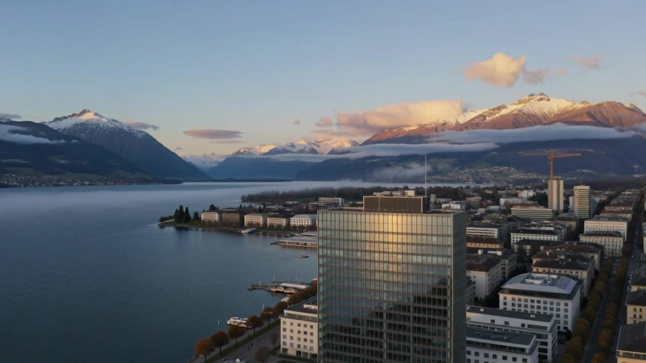 Aerial view of a glass office tower beside a Swiss lake with snow-capped mountains